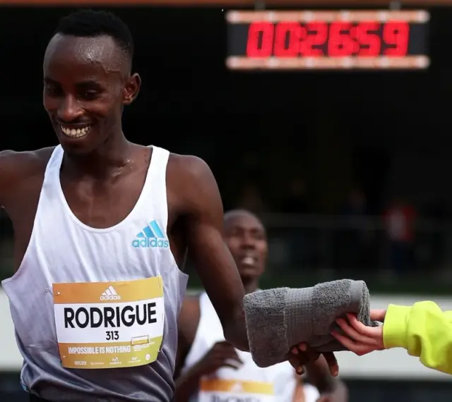 Atleta sonriente recibe toalla en una carrera de Madrid vintage. El marcador digital muestra 00:26:59.